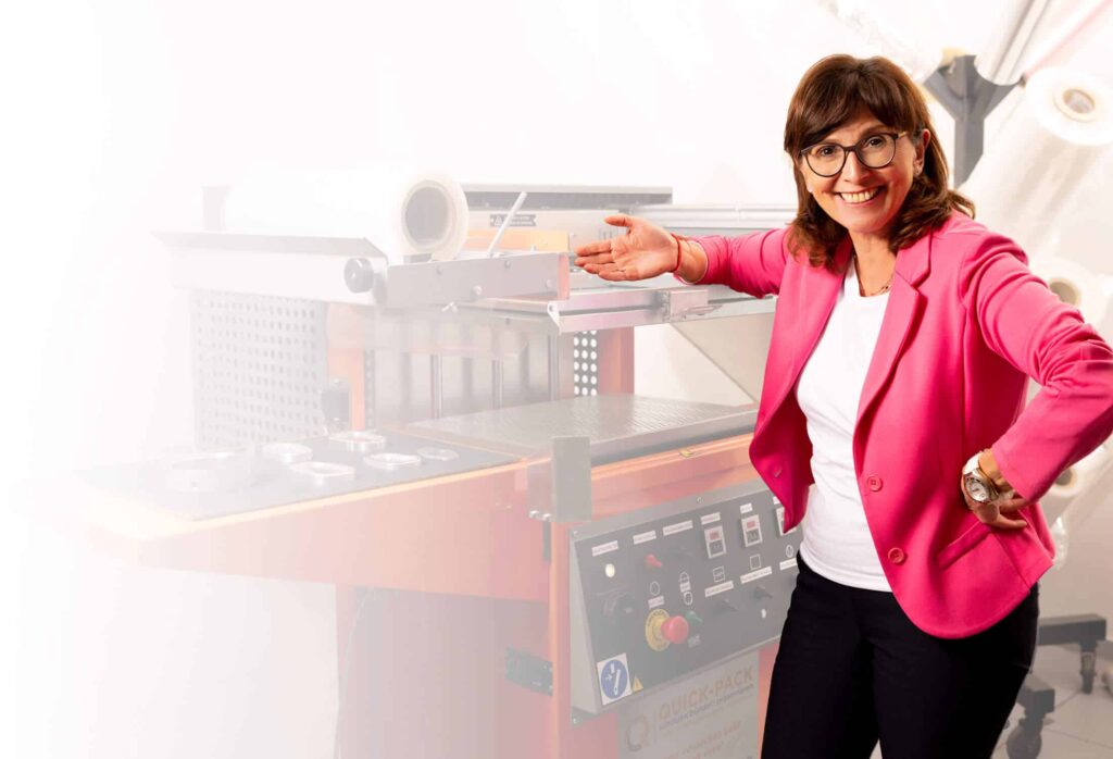 Cornelia Kehrer von QUICK-PACK in pinkem Blazer neben moderner Verpackungsmaschine mit Skinfolie im Büro.