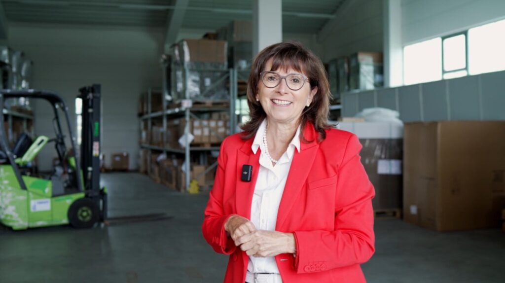 Cornelia Kehrer Geschäftsführerin von QUICK-PACK im roten Blazer steht in einem modernen Verpackungslager mit Schrumpffolie, Verpackungsmaschinen und einem grünen Forklift zwischen Metallregalen und Kartonboxen.